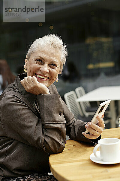 Cheerful woman holding smart phone and sitting near table at cafe