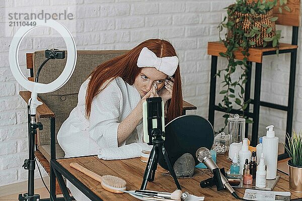 Teenage girl applying makeup and recording beauty tutorial with camera and ring light indoors.