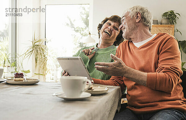 Laughing couple sitting and using tablet PC near table at home