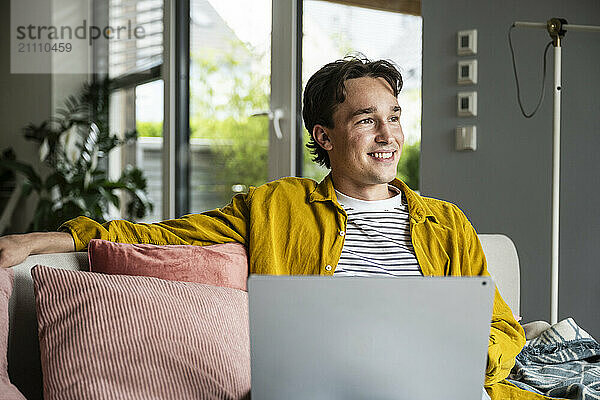 Thoughtful man sitting on sofa with laptop at home