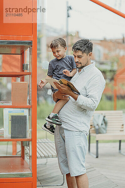 Father carrying son and reading book near book closet at park