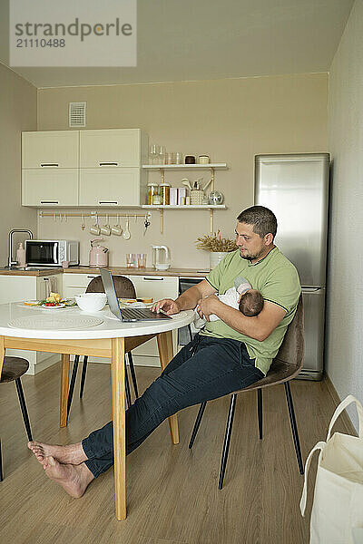 Father carrying baby and working on laptop at home