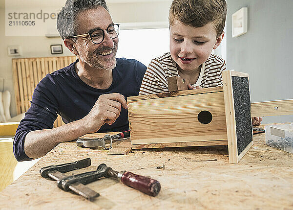 Happy grandfather making wooden birdhouse with grandson at home