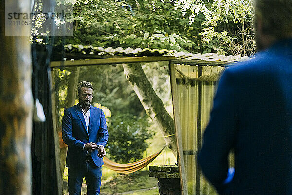 Mature businessman in blazer looking in mirror and standing at park