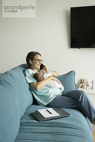 Mother holding baby and talking on smart phone sitting at home