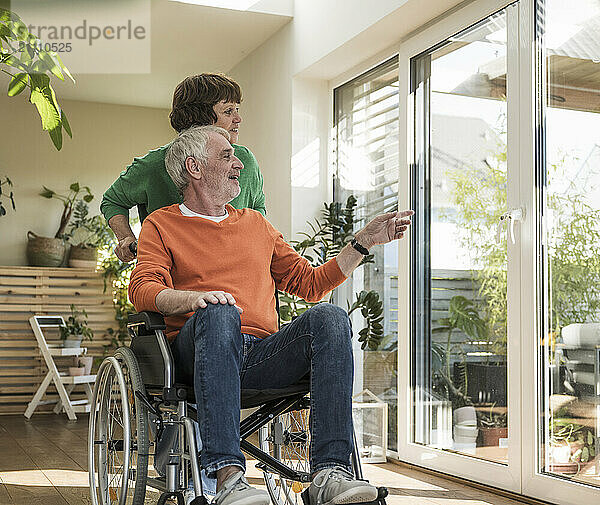 Senior woman pushing husband on wheelchair near glass door in house