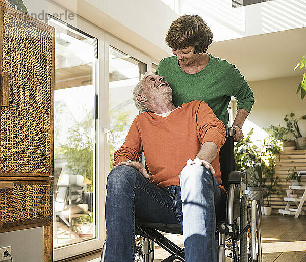 Happy senior woman pushing husband on wheelchair at home