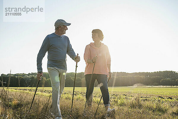 Smiling senior couple with hiking poles walking on meadow during sunny day