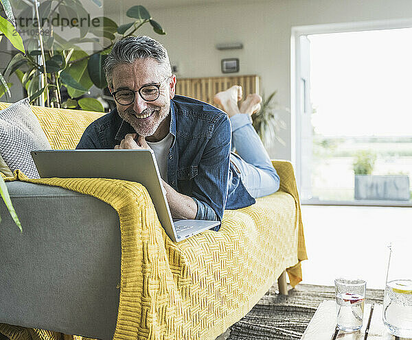 Smiling freelancer with laptop lying on sofa at home