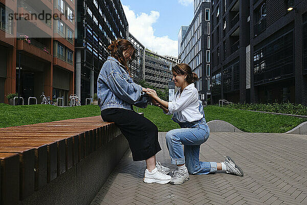 Young woman kneeling and proposing with ring to girlfriend outside building