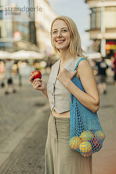 Happy woman holding apple and carrying mesh bag at street