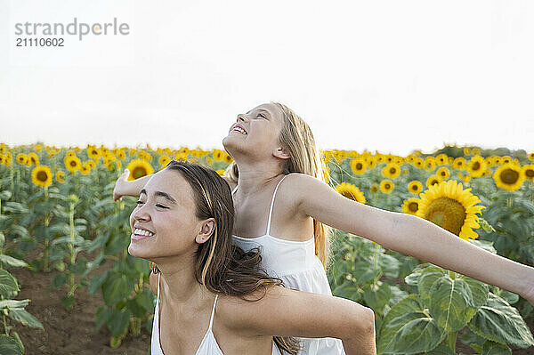 Happy woman piggybacking sister with arms outstretched in sunflower field