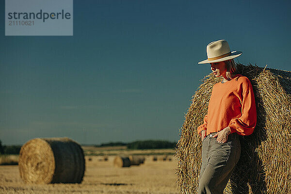 Confident woman with hands in pockets leaning on hay bale at sunny day