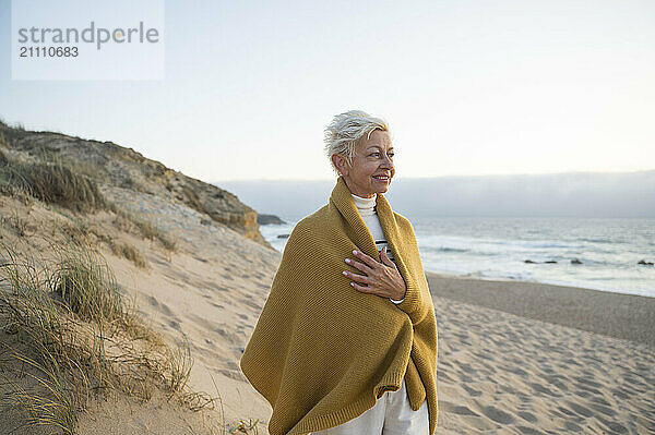 Smiling short haired woman wrapped in shawl standing on beach