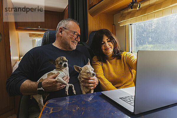 Smiling man holding dogs and using laptop with woman in motor home