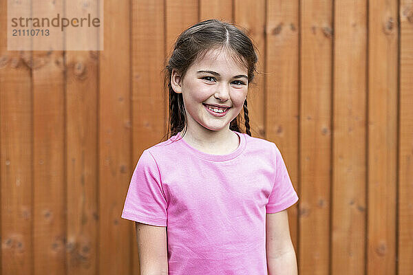 Smiling girl in pink t-shirt standing in front of wooden fence