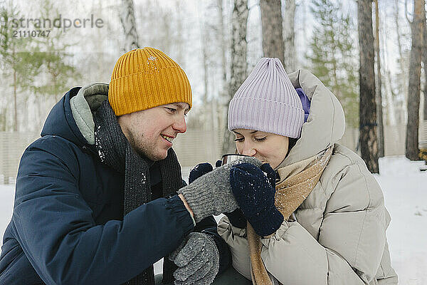 Smiling man feeding coffee to woman in warm clothes on snow