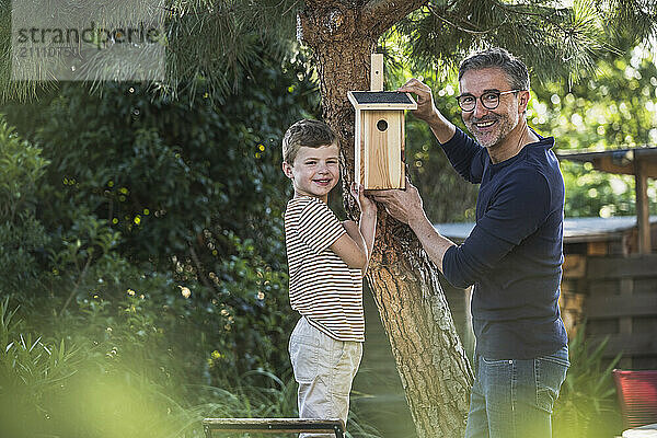 Smiling man and boy hanging birdhouse on tree in back yard