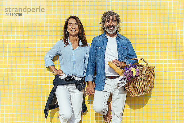 Happy senior couple with picnic basket standing in front of yellow wall