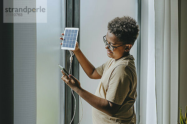 Smiling boy charging smart phone with solar panel equipment at home