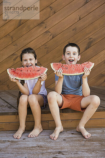 Siblings sitting and eating water melon slices at porch