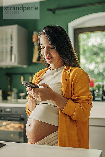 Young pregnant woman using smart phone in kitchen