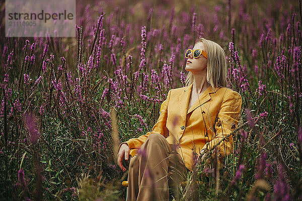 Fashionable woman sitting in yellow jacket amidst heather flowers in field