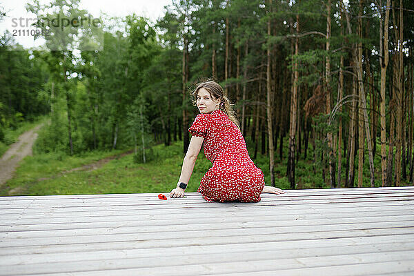 Woman looking over shoulder sitting on wooden pier near forest