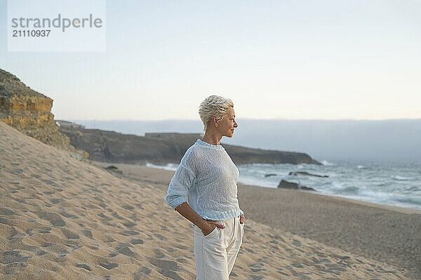 Mature short haired woman with hands in pockets standing at beach