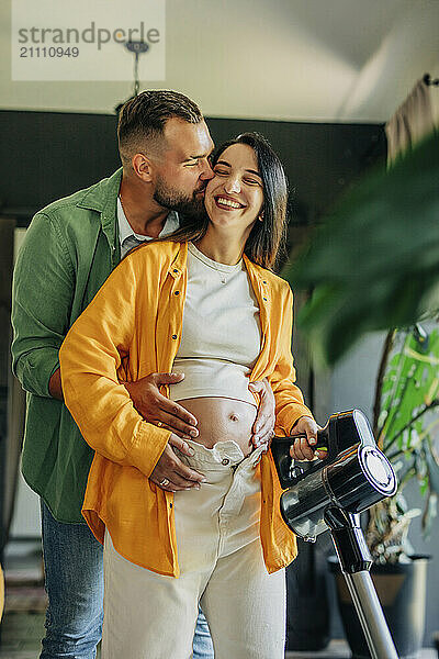 Affectionate man embracing and kissing pregnant wife vacuuming at home