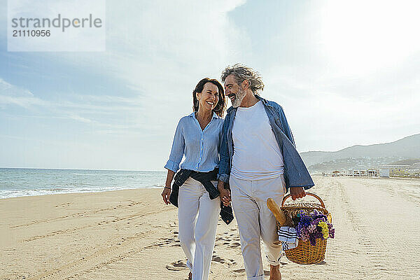 Happy couple holding hands and walking with basket at beach