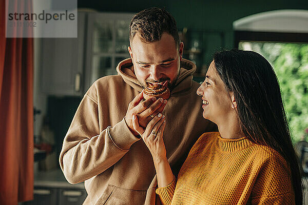 Young pregnant woman feeding cinnamon bun to husband in domestic kitchen