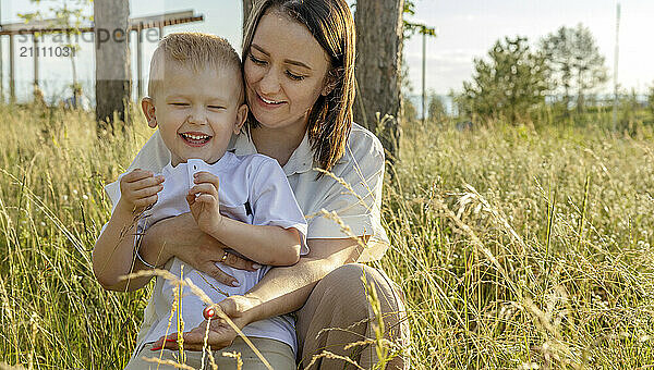 A laughing boy sits on his mother's lap in a clearing in a city park.