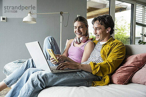 Smiling woman sharing smart phone with boyfriend sitting on sofa at home