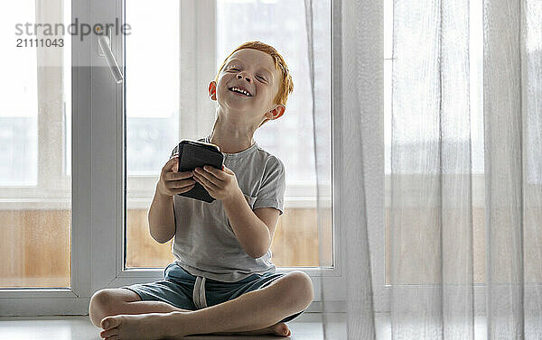 Cute boy holding smart phone and sitting on window sill at home