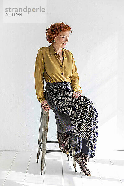 Senior redhead woman sitting on stool in front of white wall