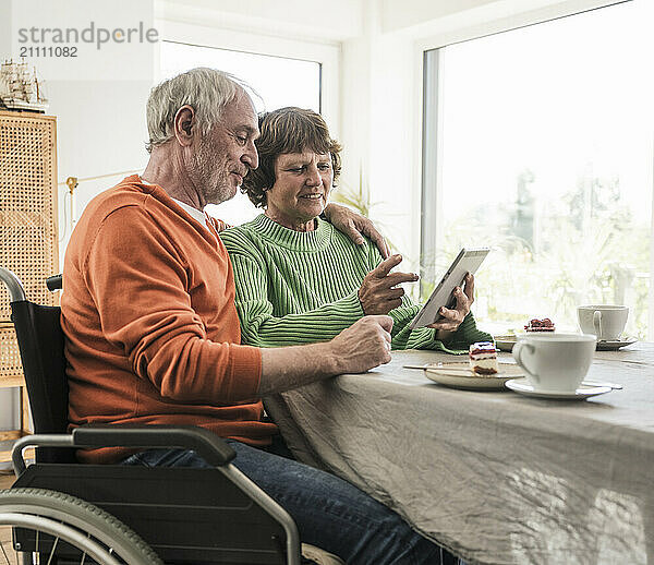 Affectionate senior man on wheelchair sitting with wife using digital PC at home