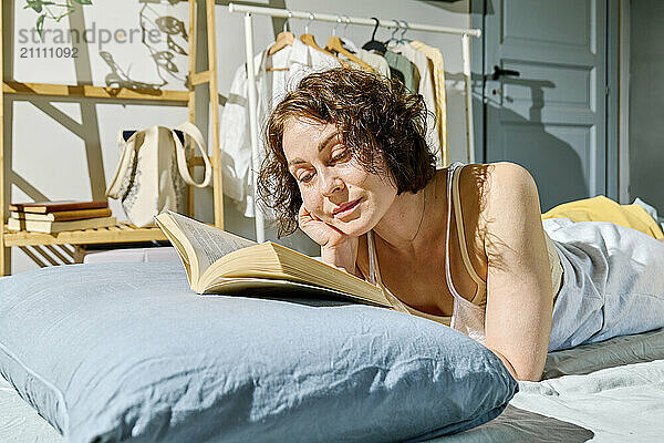 Woman with hand on chin reading book on bed at home