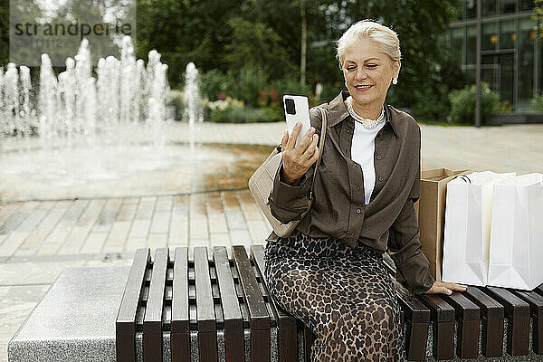 Smiling woman using smart phone and sitting on bench in street