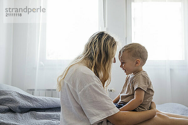 Happy boy enjoying leisure time with mother sitting on bed at home