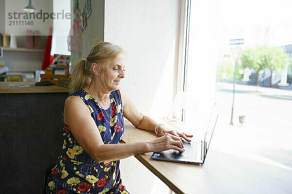 Mature businesswoman sitting near window and using laptop in office