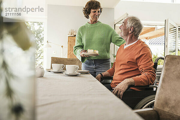 Smiling woman serving sliced cake to husband on wheelchair at home