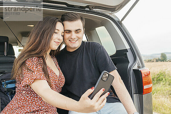 Young woman taking selfie with boyfriend in car trunk