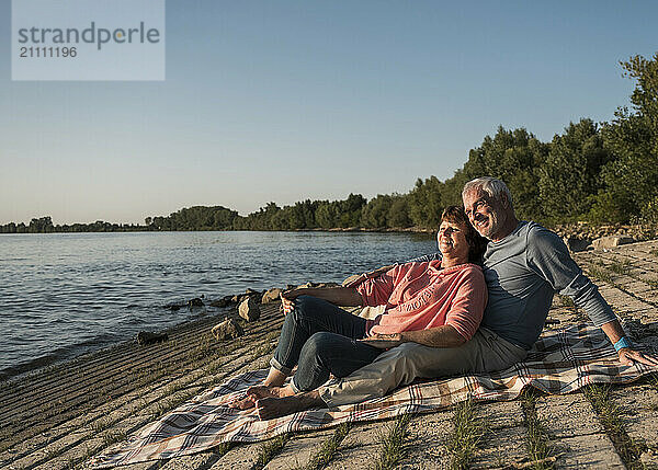 Smiling couple sitting on picnic blanket at riverbank on sunny day