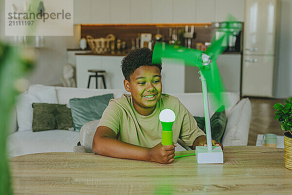 Smiling boy powering light bulb through wind turbine model in living room at home