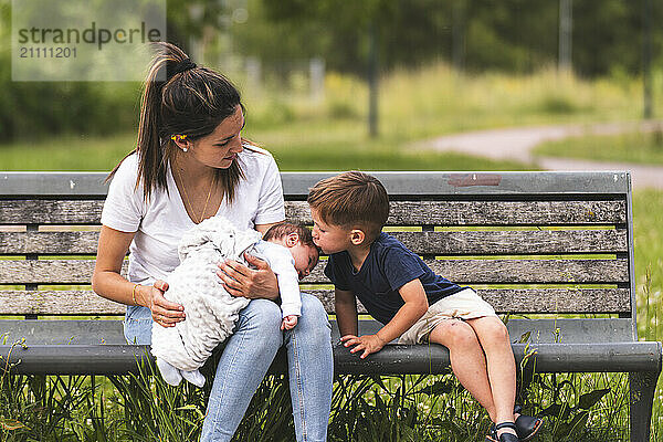 Boy kissing baby sitting with mother on bench at playground