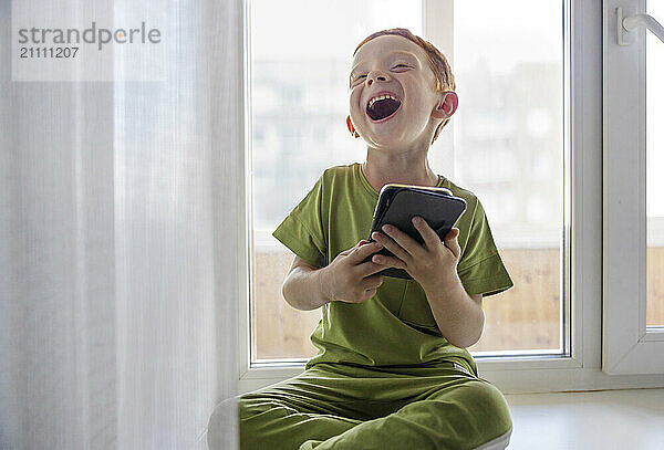 Happy boy with smart phone sitting on window sill