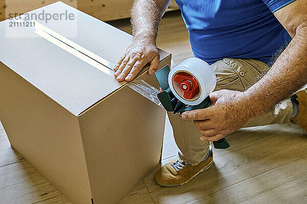 Senior man kneeling and taping cardboard box in new apartment