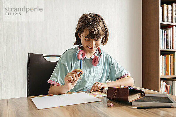 Smiling girl holding pencil and sitting near table at home