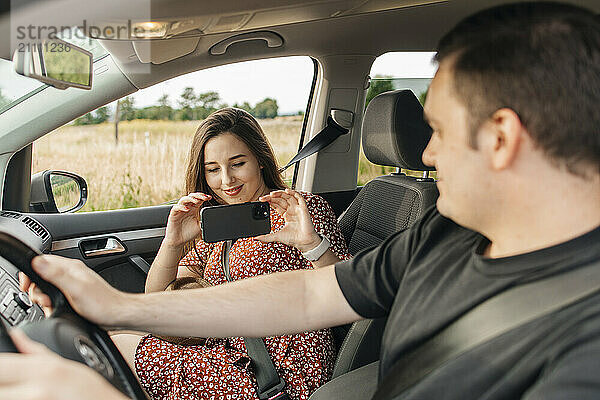 Young woman photographing boyfriend through smart phone in car